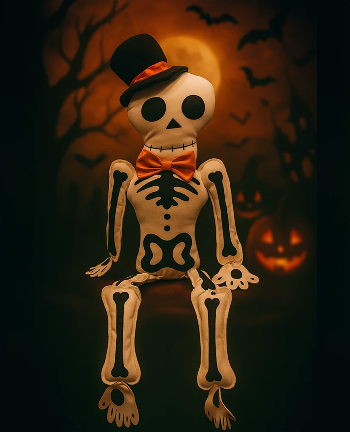 A Halloween skeleton doll wearing a black top hat with an orange band and an orange bow tie, posed against a spooky dark background with bats, a full moon, and a jack-o'-lantern.