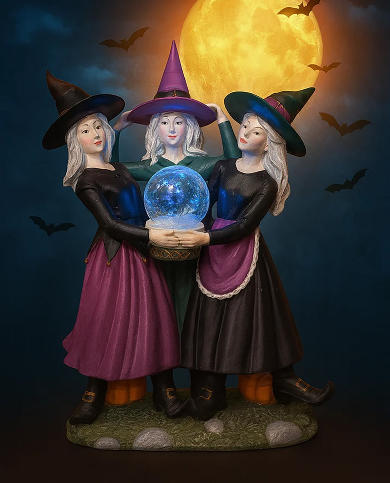 A Halloween-themed figurine of three witches holding a glowing blue crystal ball, set against a dark night sky with a large full moon and flying bats.