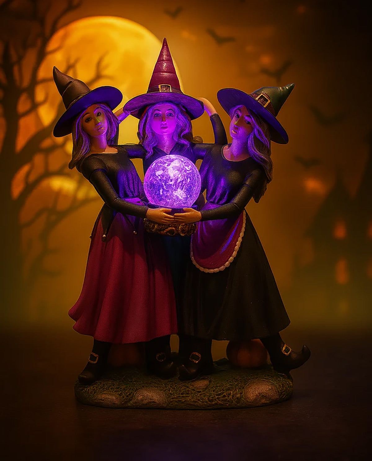 A Halloween figurine of three witches holding a glowing purple crystal ball, set against a dark background with a large full moon and silhouetted trees.