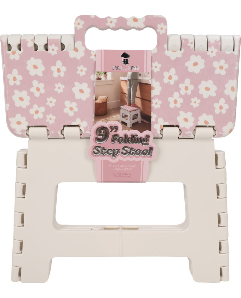 A 9-inch folding step stool shown folded flat with its pink floral retail packaging attached. The top panel has a pink background with a white daisy floral pattern, and the lower folded frame is off-white plastic.