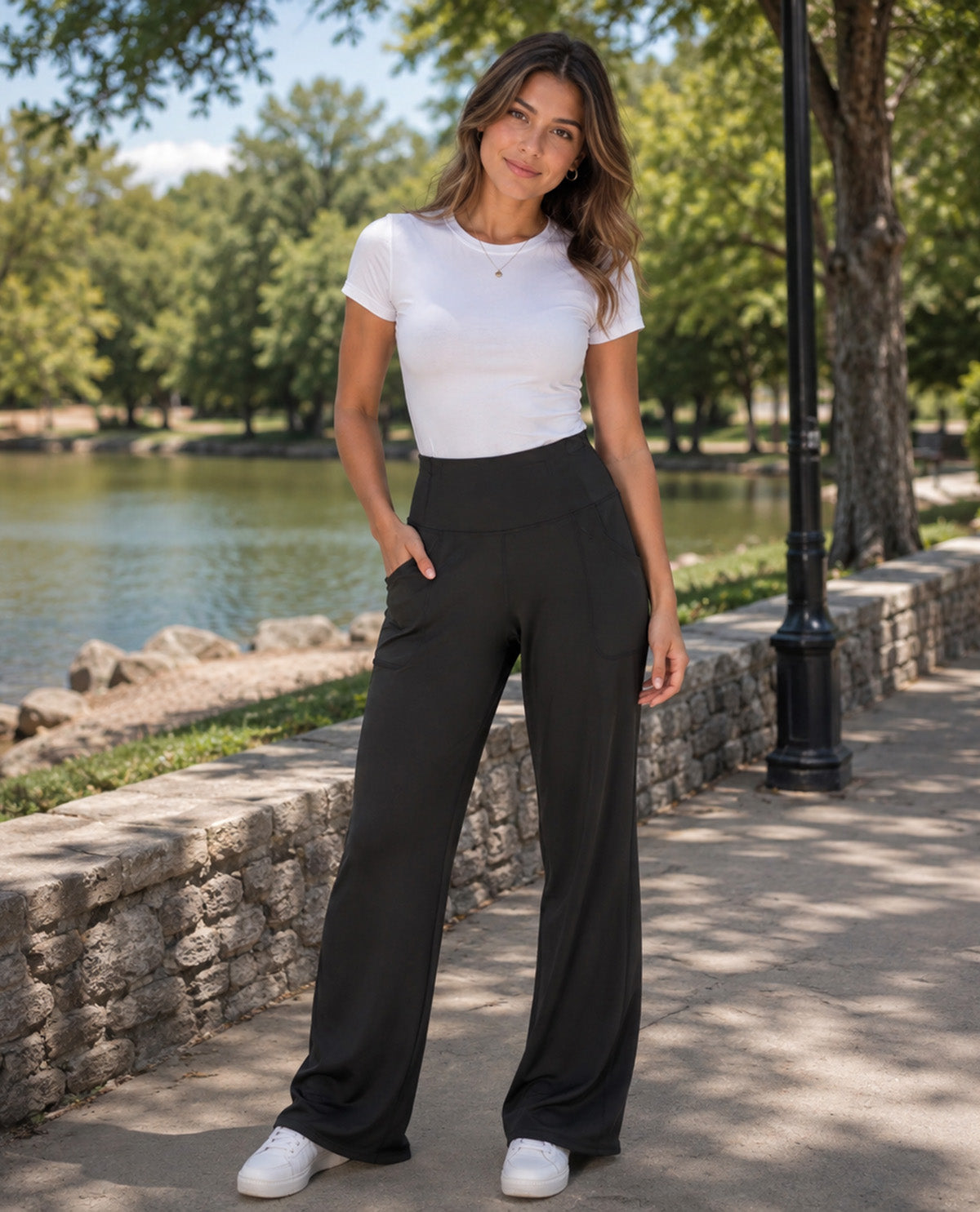 A woman stands outdoors by a lake wearing a fitted white short-sleeve top, black wide-leg pants, and white sneakers.