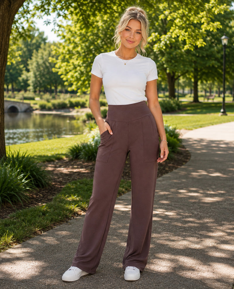 A woman stands on a paved path in a sunny park, wearing a white short-sleeve crewneck t-shirt and high-waisted, wide-leg taupe pants with white sneakers.