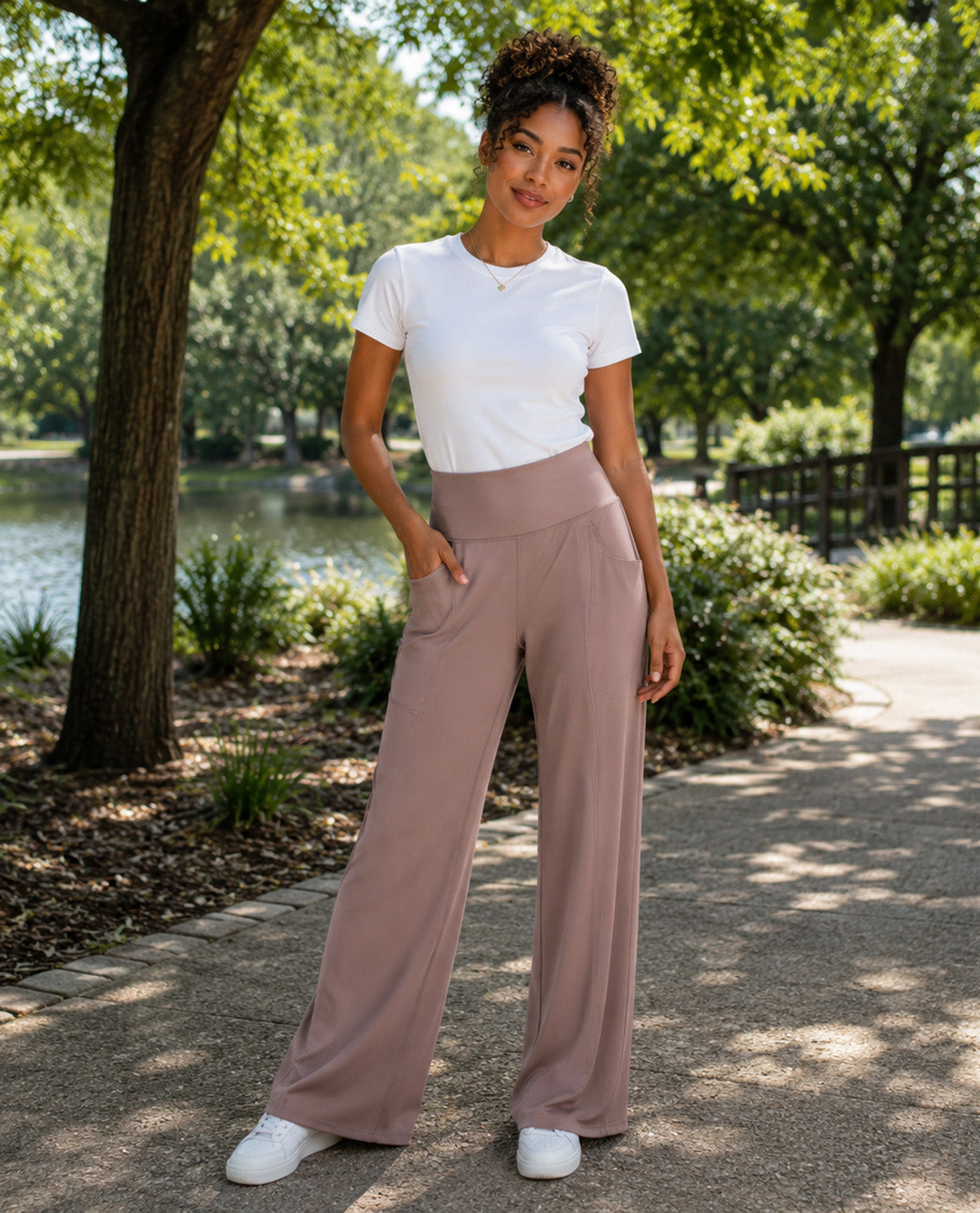 A woman stands on a tree-lined path next to a lake, wearing a white short-sleeve crewneck t-shirt and high-waisted, wide-leg taupe pants with white sneakers.
