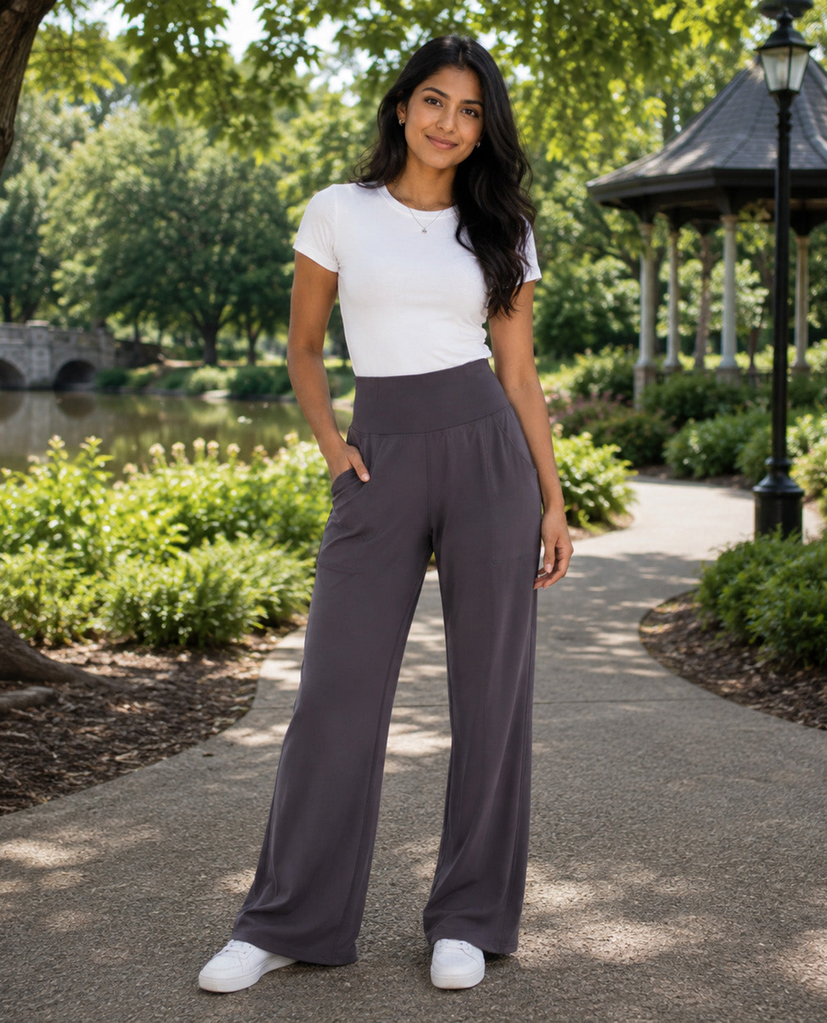 A woman stands on a park pathway wearing high-waisted, wide-leg charcoal gray pants, a white short-sleeve crewneck top, and white sneakers. The background features a lush green park with trees, a pond, and a gazebo.