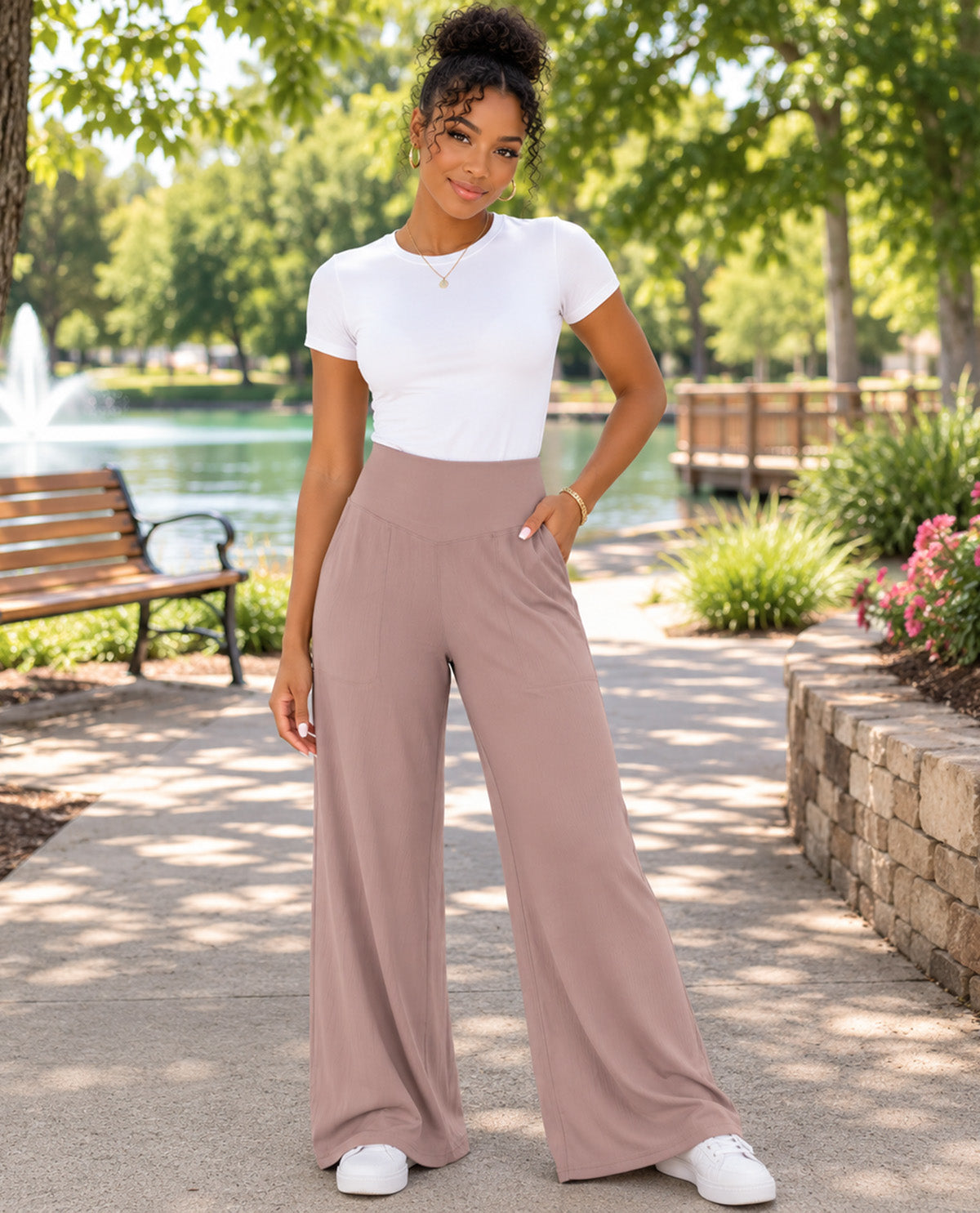 A woman stands on a paved path in a sunny park wearing a white short-sleeve crewneck top and high-waisted, wide-leg dusty rose pants, paired with white sneakers.