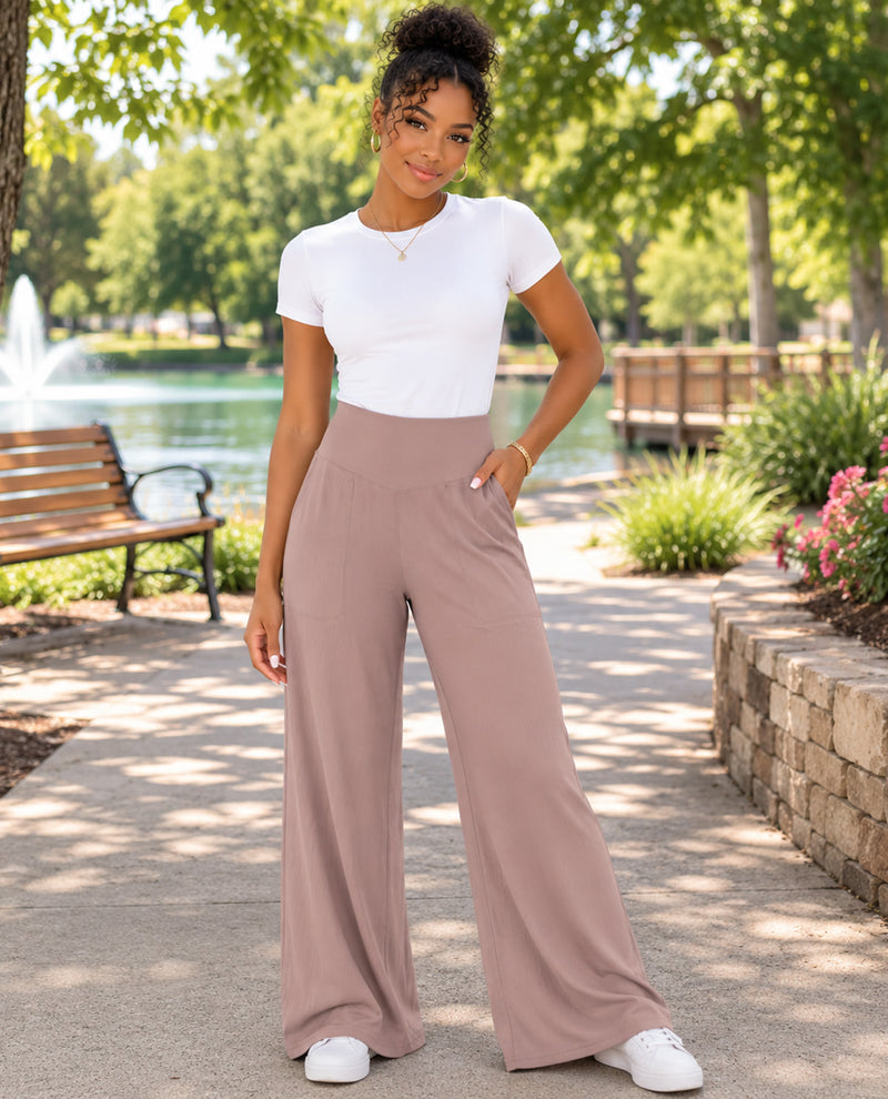 A woman stands on a paved path in a sunny park wearing a white short-sleeve crewneck top and high-waisted, wide-leg dusty rose pants, paired with white sneakers.