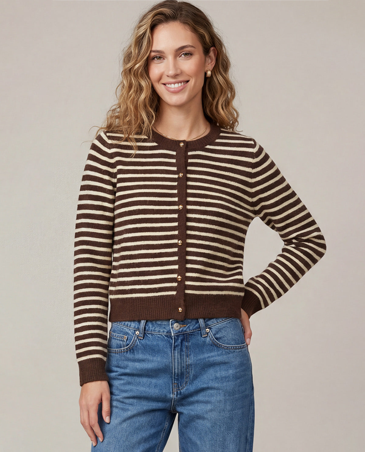 A woman wearing a brown and beige horizontally striped button-up cardigan sweater with blue jeans, standing against a solid light gray studio background.