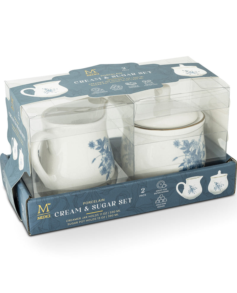 A two-piece cream and sugar set in its original retail packaging. The set includes a white ceramic mug with a blue floral pattern and a matching white creamer with a lid, both decorated with gold trim.