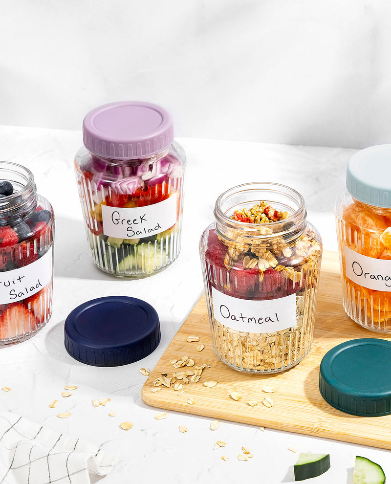 Several clear glass jars with colorful screw-on lids are arranged on a white marble-like surface. One open jar labeled "Oatmeal" sits on a wooden board, with oats and dried fruit visible inside.