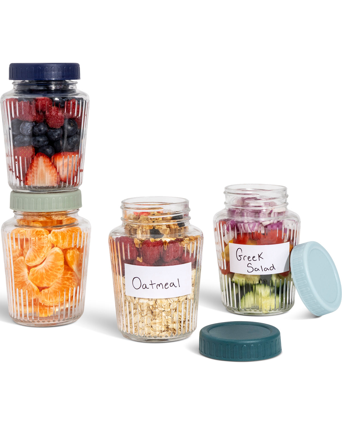 A set of clear ribbed glass jars with screw lids, shown on a white background. Two stacked jars on the left contain fruit, one open jar in the center contains oatmeal with a handwritten label reading 'Oatmeal', and one open jar on the right contains Greek salad with a handwritten label reading 'Greek Salad'.