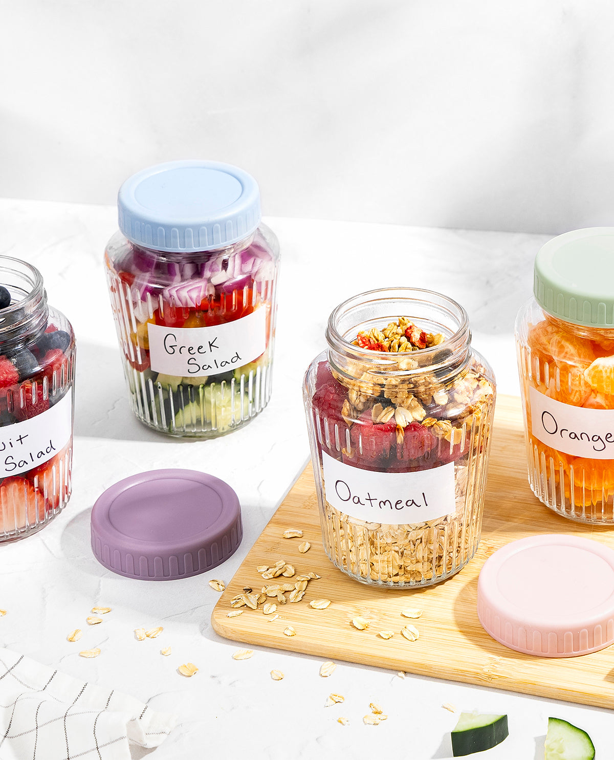 Four clear ribbed storage jars with pastel-colored screw-on lids and handwritten paper labels, arranged on a white surface with a wooden board. The jars contain colorful foods including salad, Greek salad, oatmeal, and orange slices.