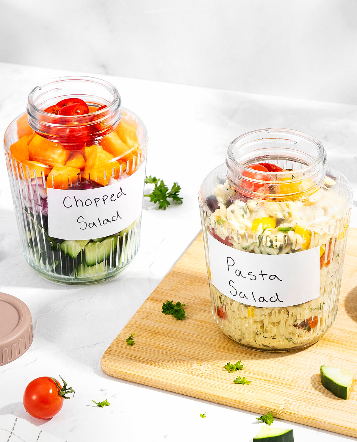 Two clear glass jars filled with salads, labeled 'Chopped Salad' and 'Pasta Salad', arranged on a white surface with a wooden cutting board and fresh tomatoes and cucumber pieces.