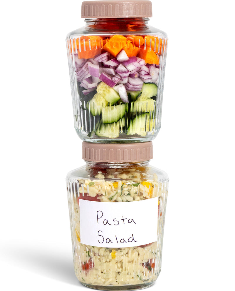 Two clear plastic food storage jars with beige screw-top lids stacked vertically on a white background. The top jar contains layered chopped vegetables including carrots, red onion, and cucumbers, while the bottom jar has a handwritten label reading 'Pasta Salad' and contains mixed pasta salad.