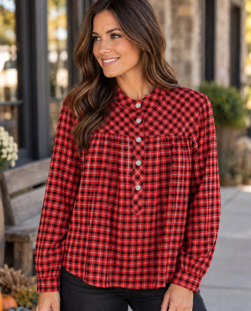 A woman wearing a red and black buffalo plaid long-sleeve button-up shirt, standing outdoors in front of a blurred building background.