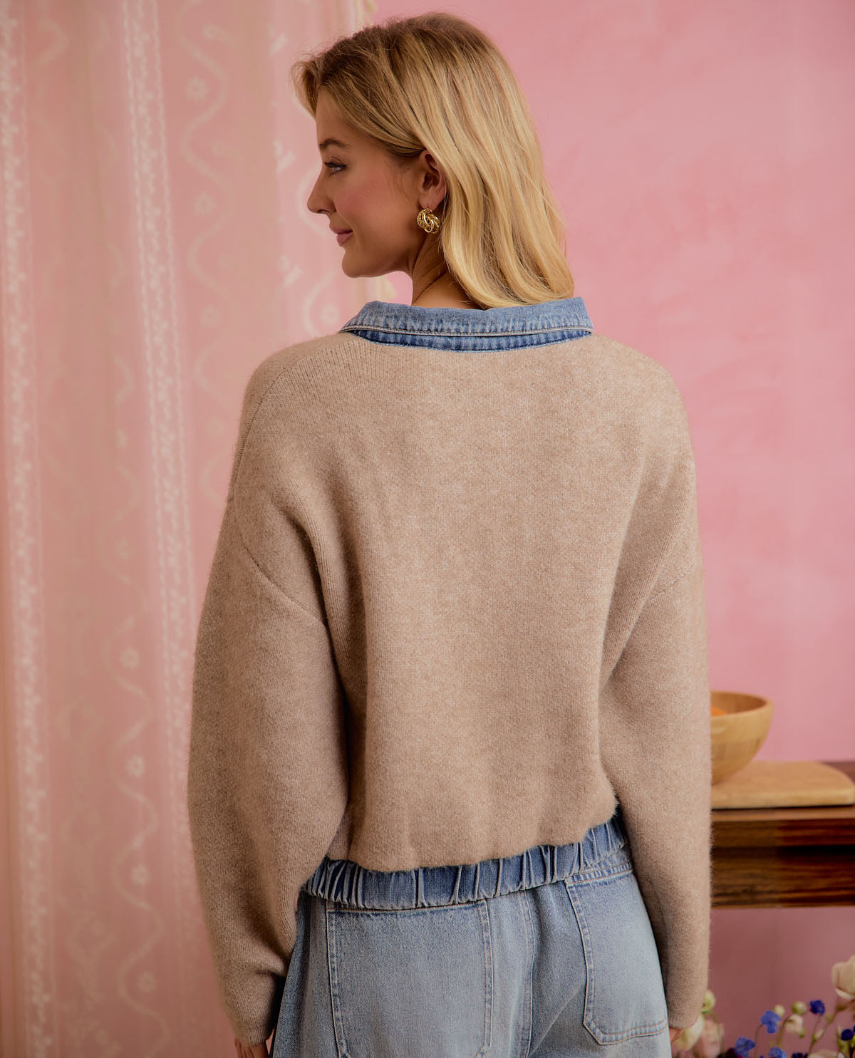 A woman shown from the back wearing a beige knit sweater with a blue denim collar and matching blue denim waistband, standing against a pink wall.
