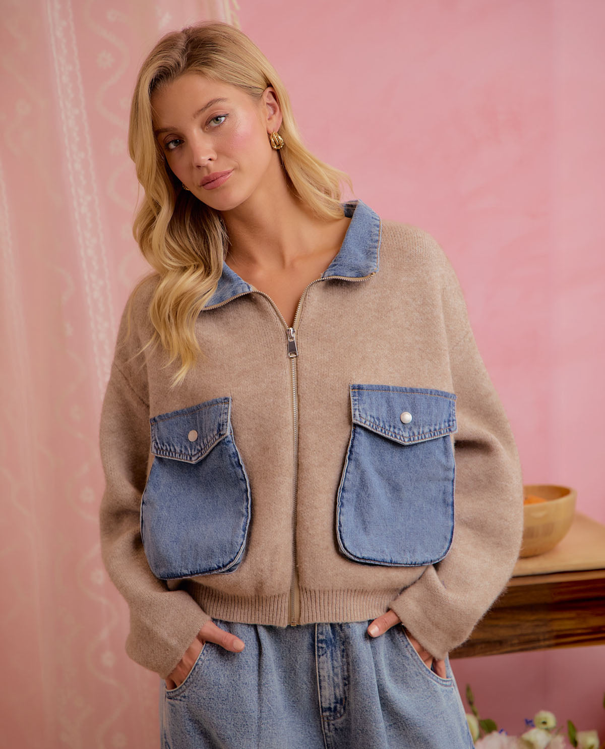 A woman wearing a beige knit zip-up jacket with blue denim pockets and collar, paired with light-wash jeans, posed against a pink patterned backdrop.