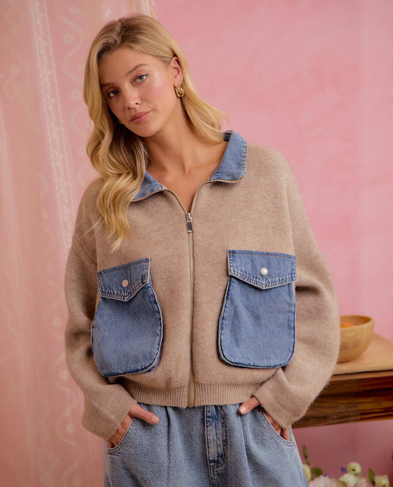 A woman wearing a beige knit zip-up jacket with blue denim pockets and collar, paired with light-wash jeans, posed against a pink patterned backdrop.