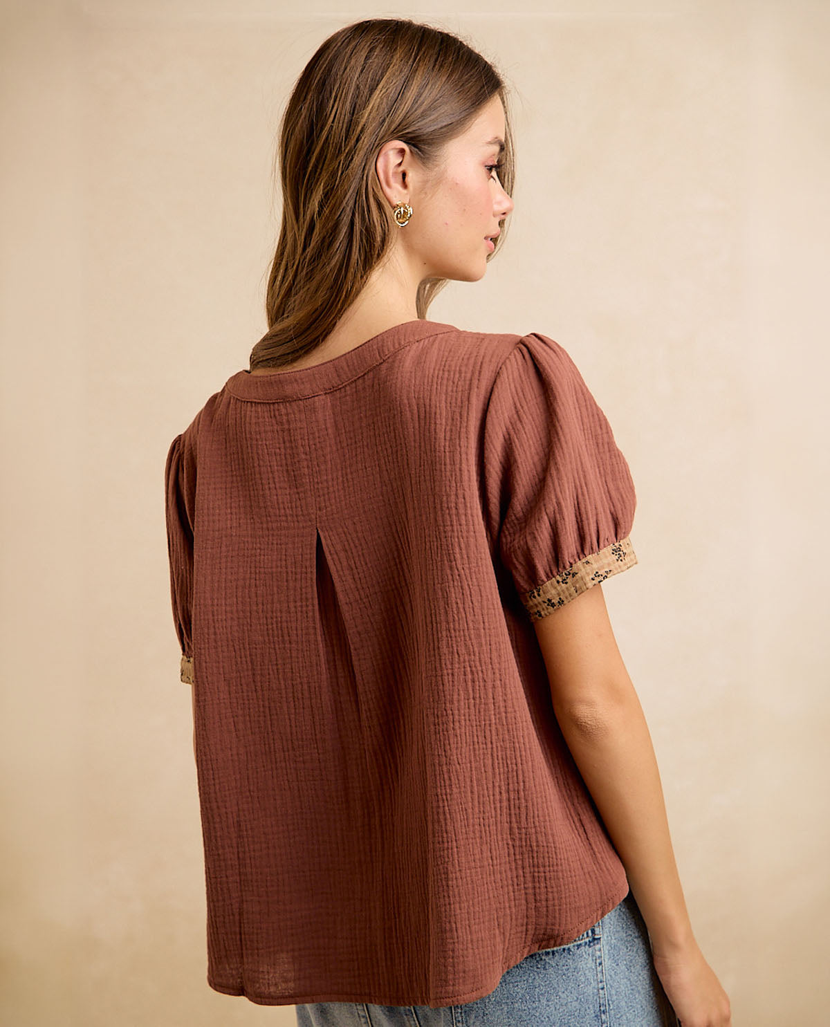 A woman shown from the back wearing a textured rust-brown short-sleeve blouse with puff sleeves and lace trim, paired with blue jeans against a neutral beige background.