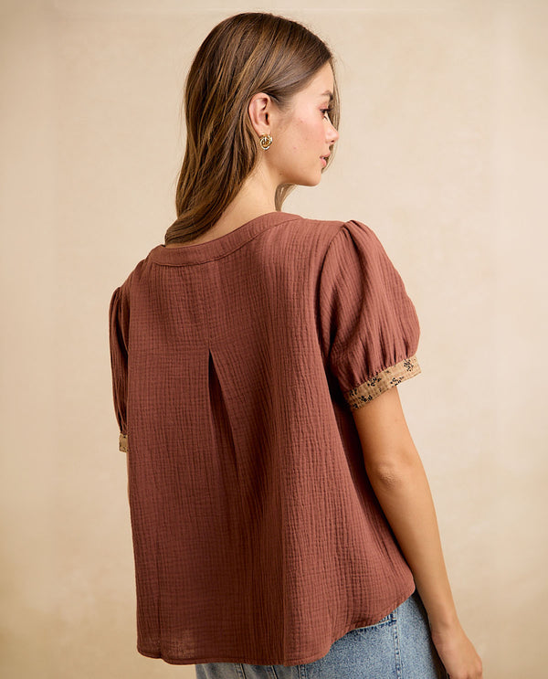 A woman shown from the back wearing a textured rust-brown short-sleeve blouse with puff sleeves and lace trim, paired with blue jeans against a neutral beige background.