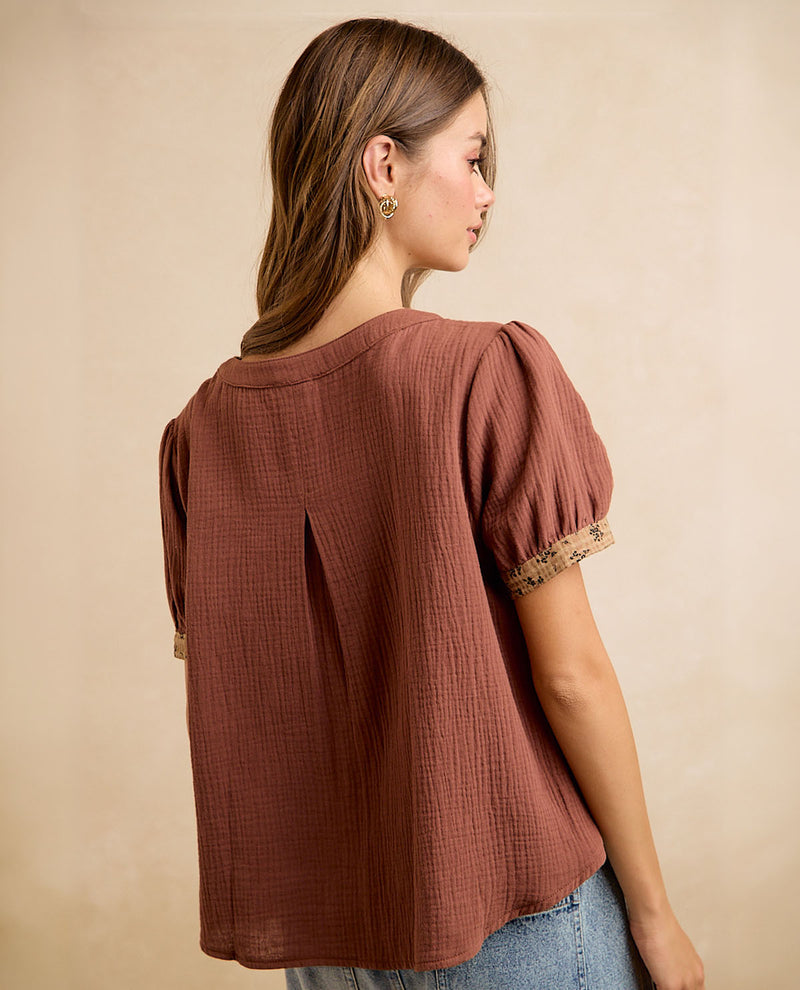 A woman shown from the back wearing a textured rust-brown short-sleeve blouse with puff sleeves and lace trim, paired with blue jeans against a neutral beige background.