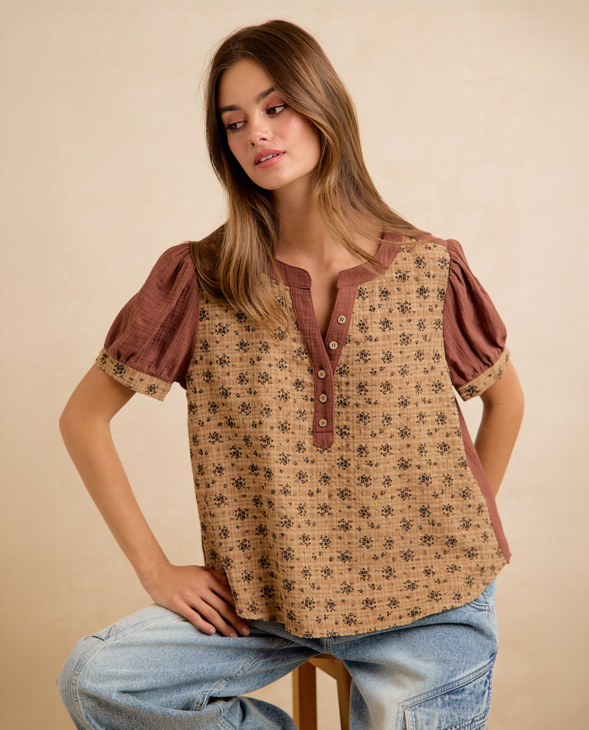 A woman wearing a short-sleeve button-front top with a brown and beige floral pattern, seated against a plain beige studio background.