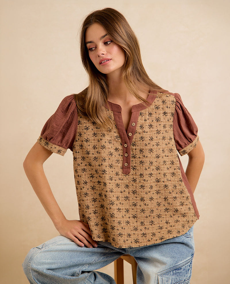 A woman wearing a short-sleeve button-front top with a brown and beige floral pattern, seated against a plain beige studio background.