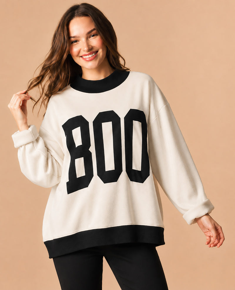 A woman wearing a cream-colored crewneck sweatshirt with black trim and large black 'BOO' lettering on the front, paired with black pants, set against a solid beige background.
