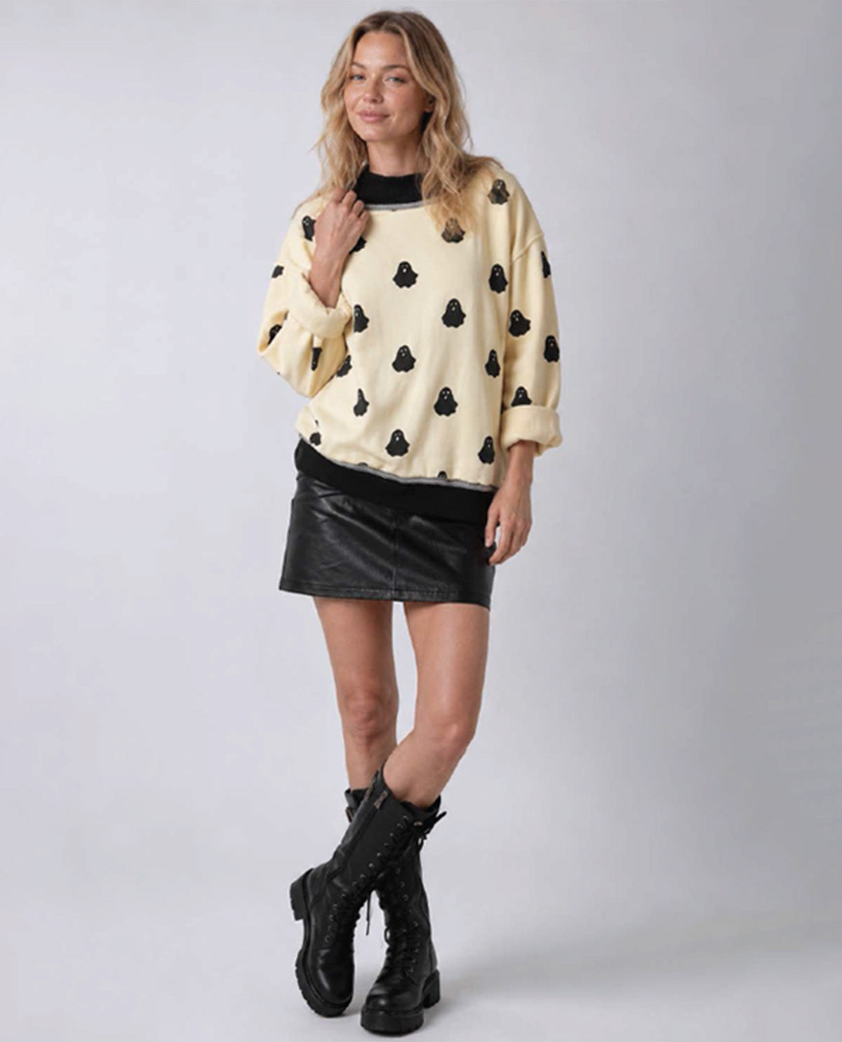 A woman modeling a cream-colored long-sleeve sweatshirt with black playing card suits, paired with a black faux-leather mini skirt and black combat boots, against a light gray studio background.
