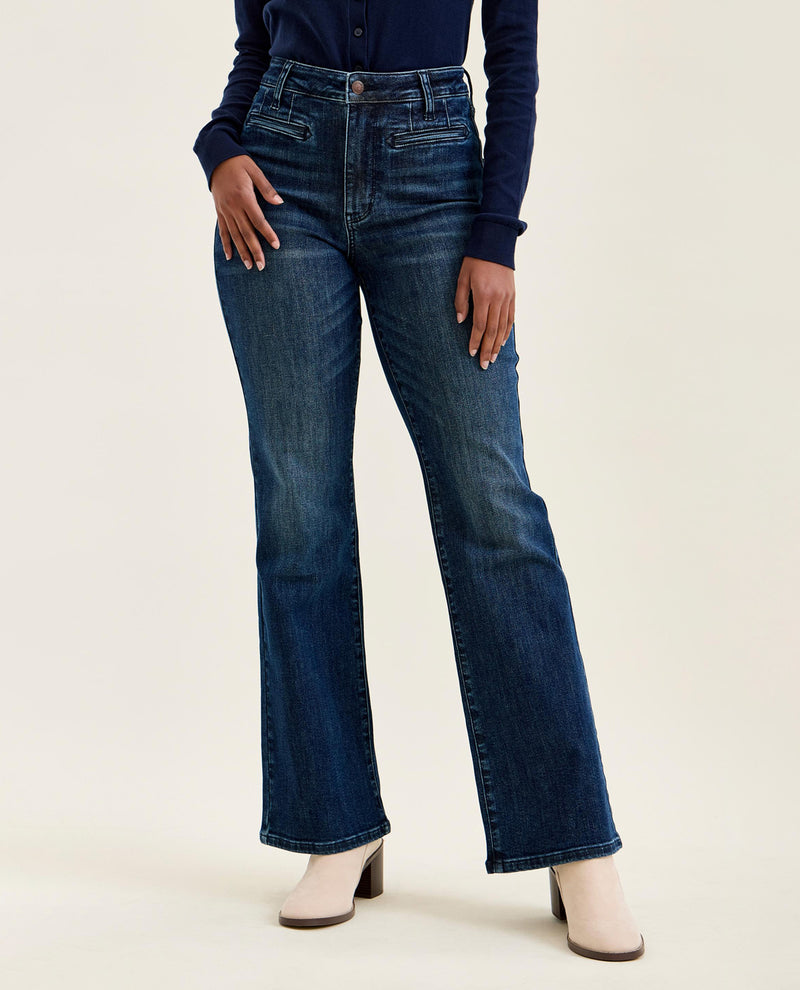 A model wearing dark blue high-waisted flare jeans, a navy long-sleeve top, and beige heeled mules against a plain off-white studio background.