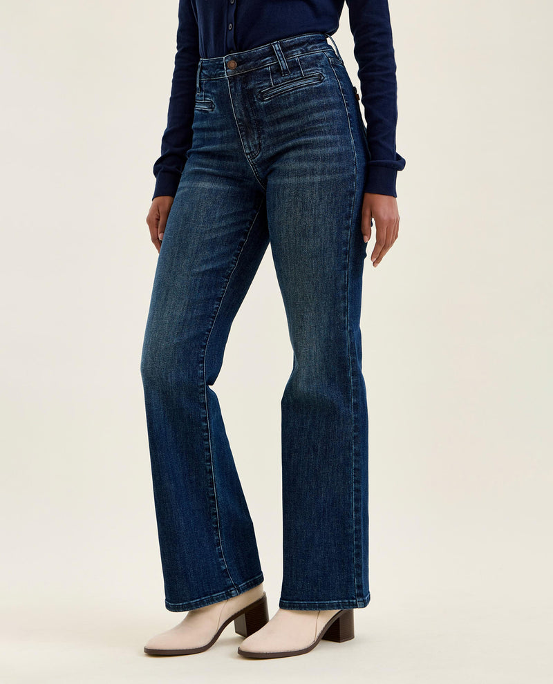 A model wearing dark blue high-waisted flare jeans, a navy long-sleeve top, and beige heeled mules against a plain off-white studio background.