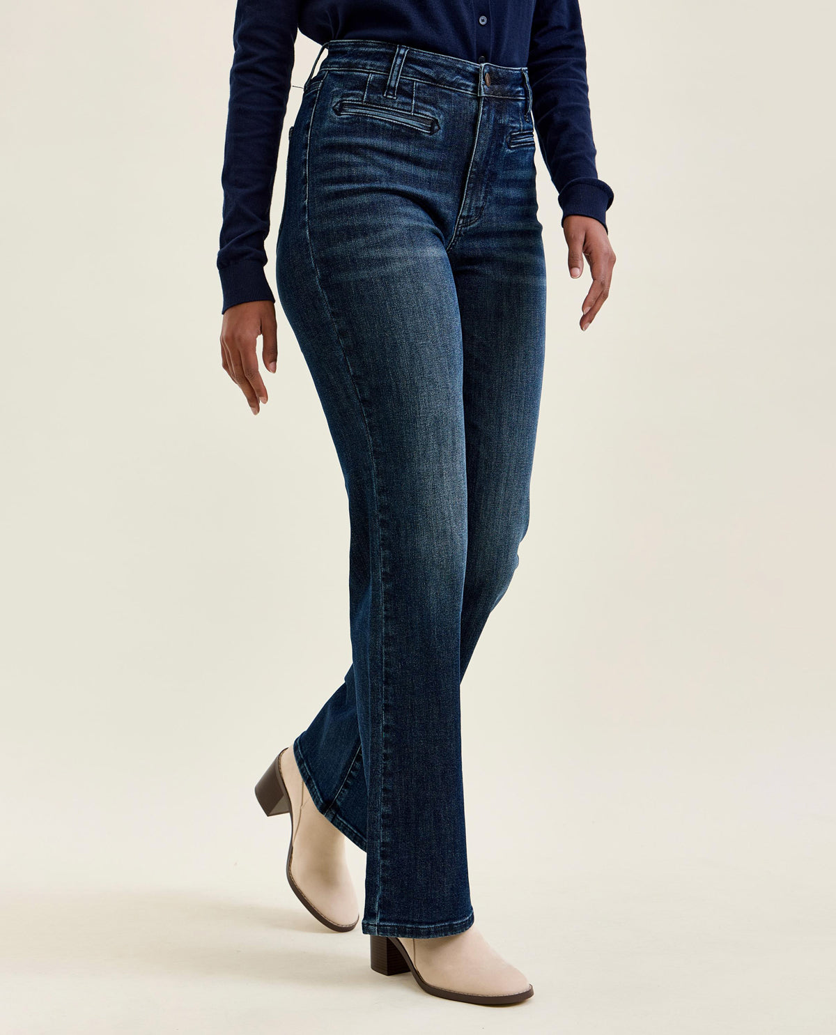 A model wearing dark blue high-waisted flare jeans, a navy long-sleeve top, and beige heeled ankle boots, shown from the waist down against a light beige studio background.