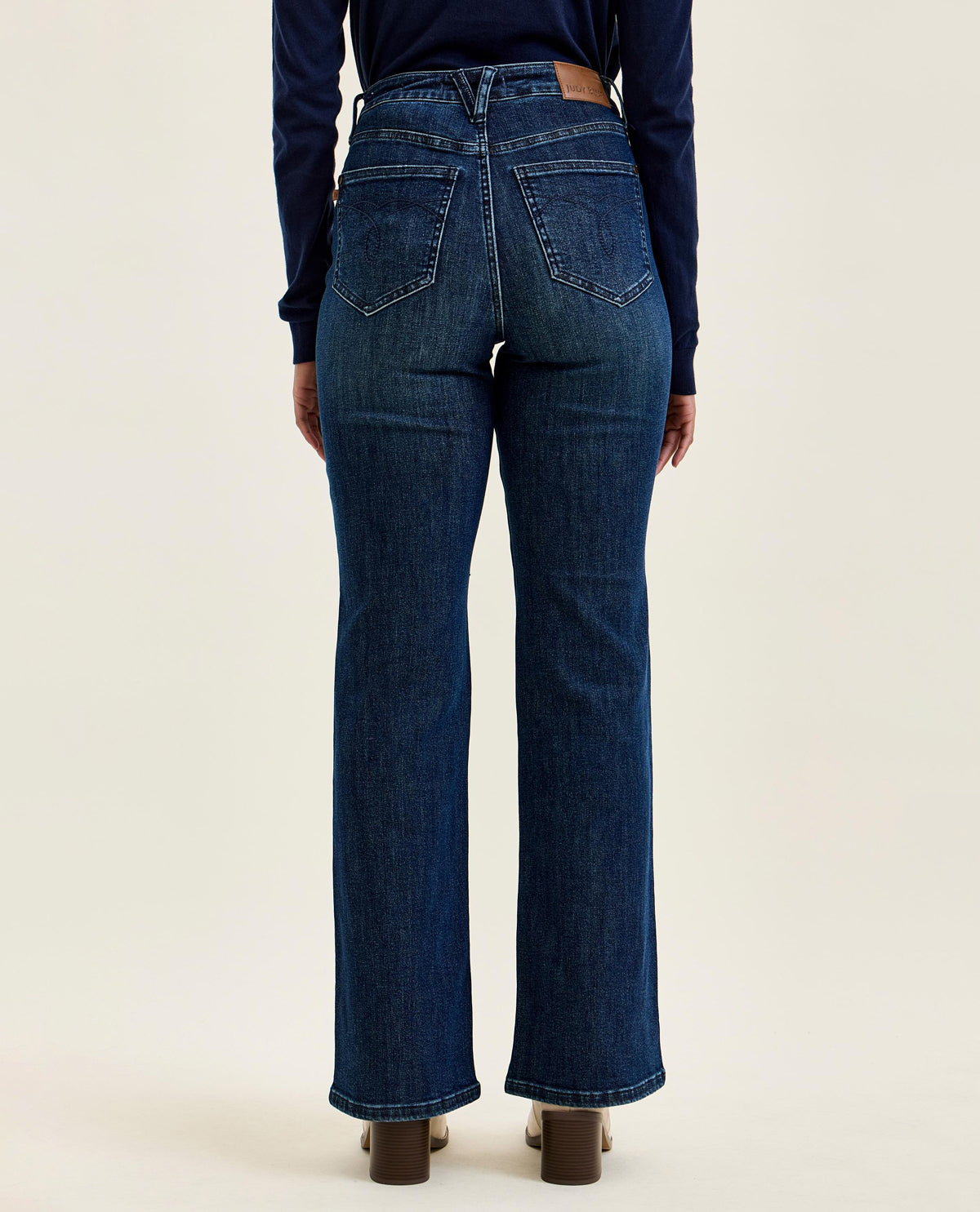 Rear view of a model wearing dark blue high-waisted flare jeans with a brown leather patch at the back waist, paired with a navy long-sleeve top and brown block heels, against a light beige studio background.