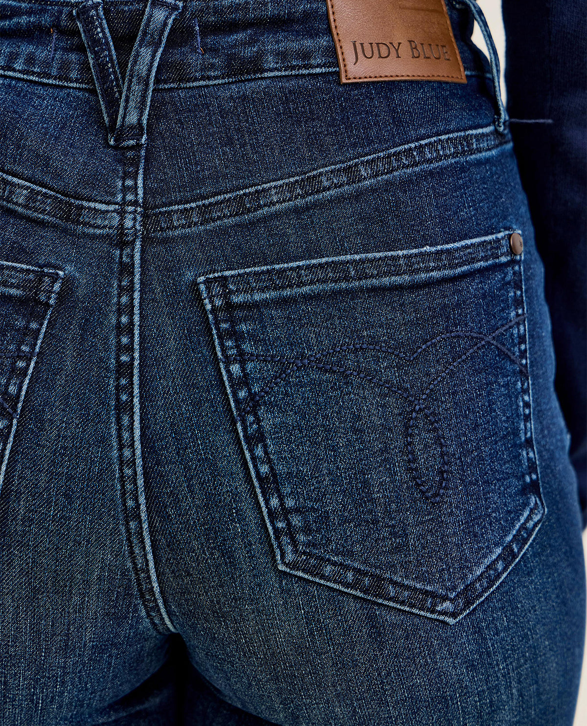 Close-up of the back of dark blue denim jeans showing the rear pocket, stitching, and a brown leather patch with the brand name JUDY BUE.