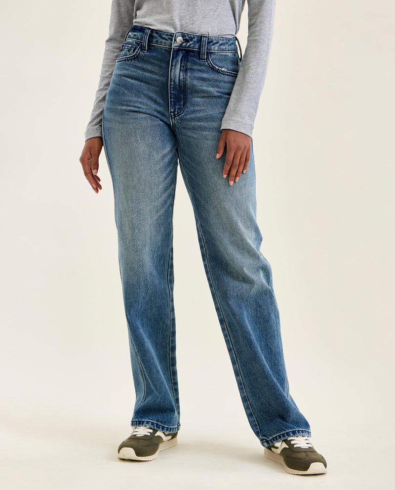 A person wearing a pair of high-waisted, straight-leg blue denim jeans with a gray long-sleeve top and gray sneakers, standing against a plain light beige studio background.