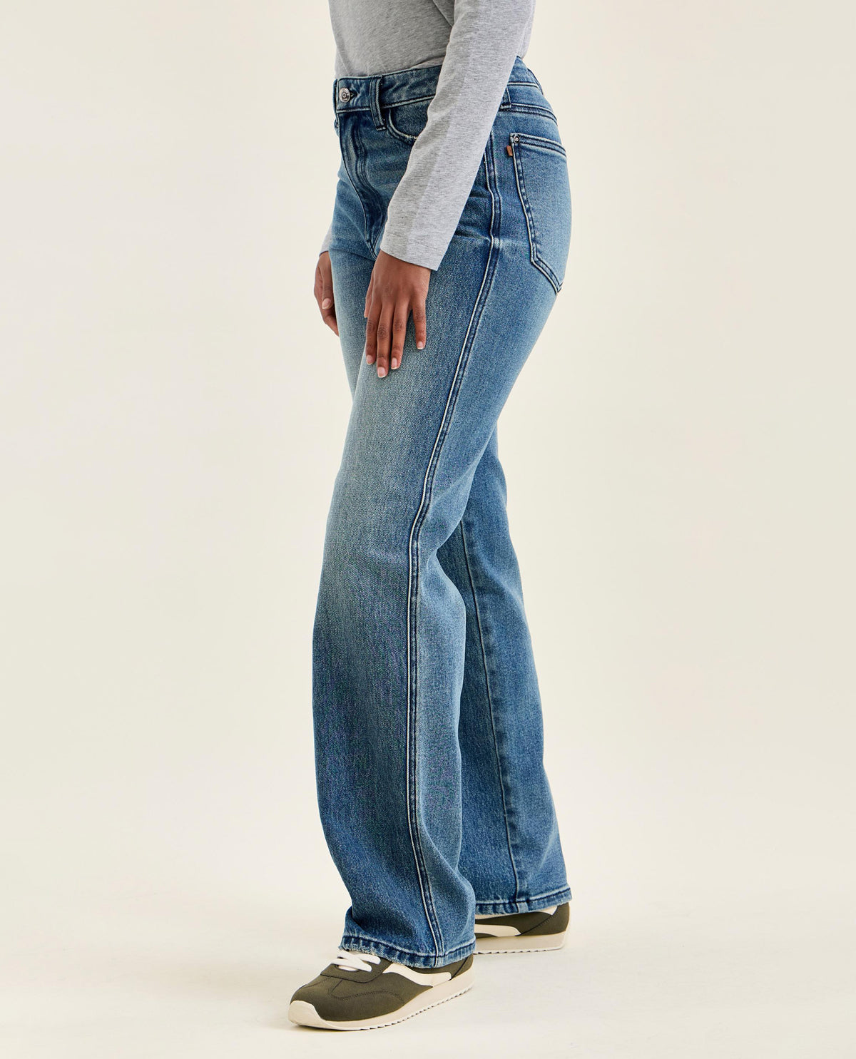 A side profile view of a person wearing high-waisted, wide-leg blue jeans, a light gray long-sleeve top, and dark gray sneakers against a plain off-white studio background.
