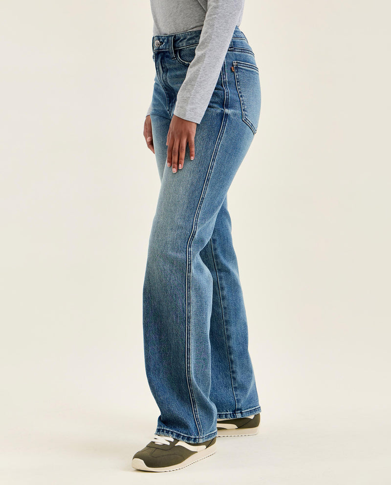 A side profile view of a person wearing high-waisted, wide-leg blue jeans, a light gray long-sleeve top, and dark gray sneakers against a plain off-white studio background.