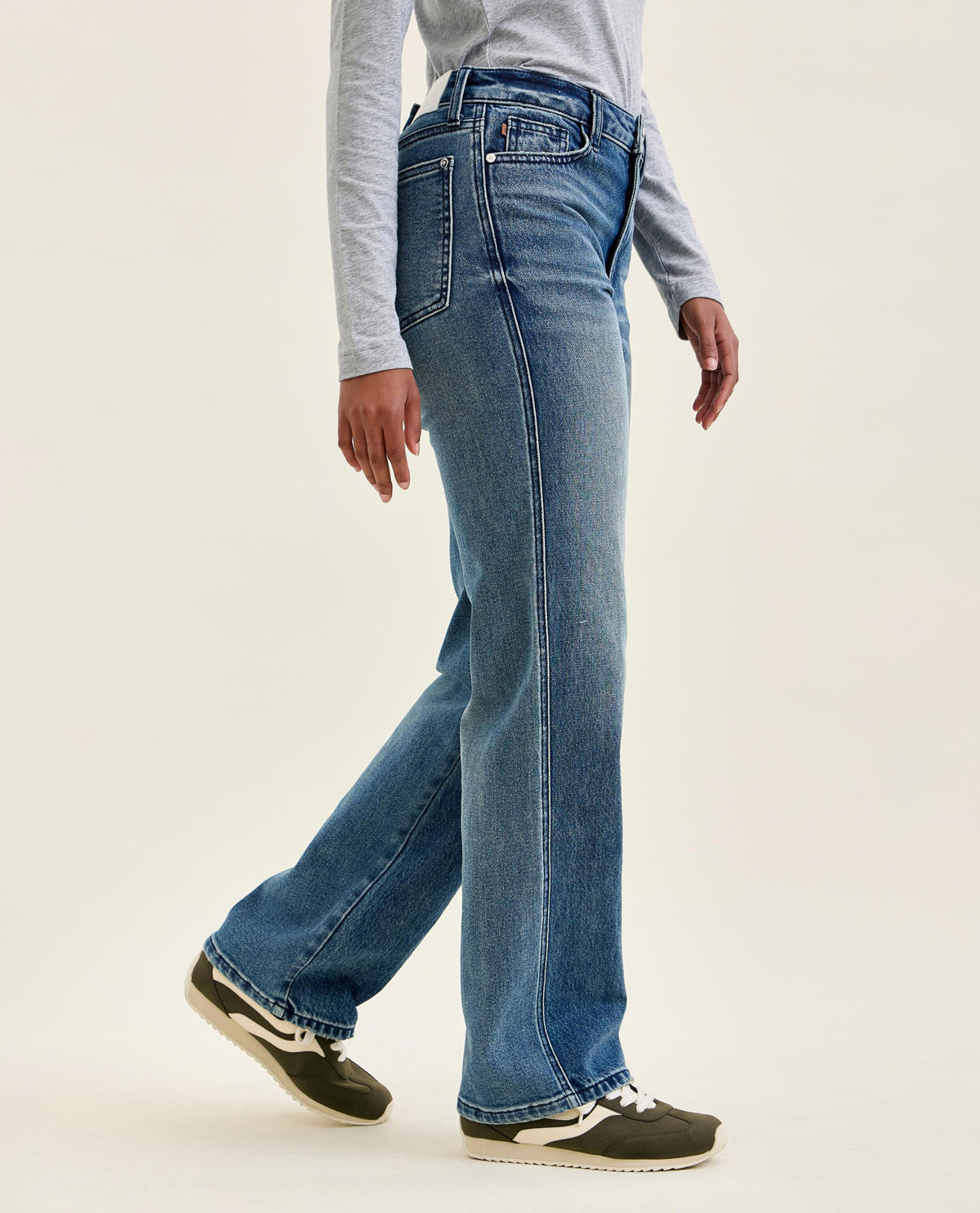 A person wearing medium-wash blue wide-leg jeans, a light gray long-sleeve top, and dark green sneakers, shown from the waist down against a plain off-white studio background.