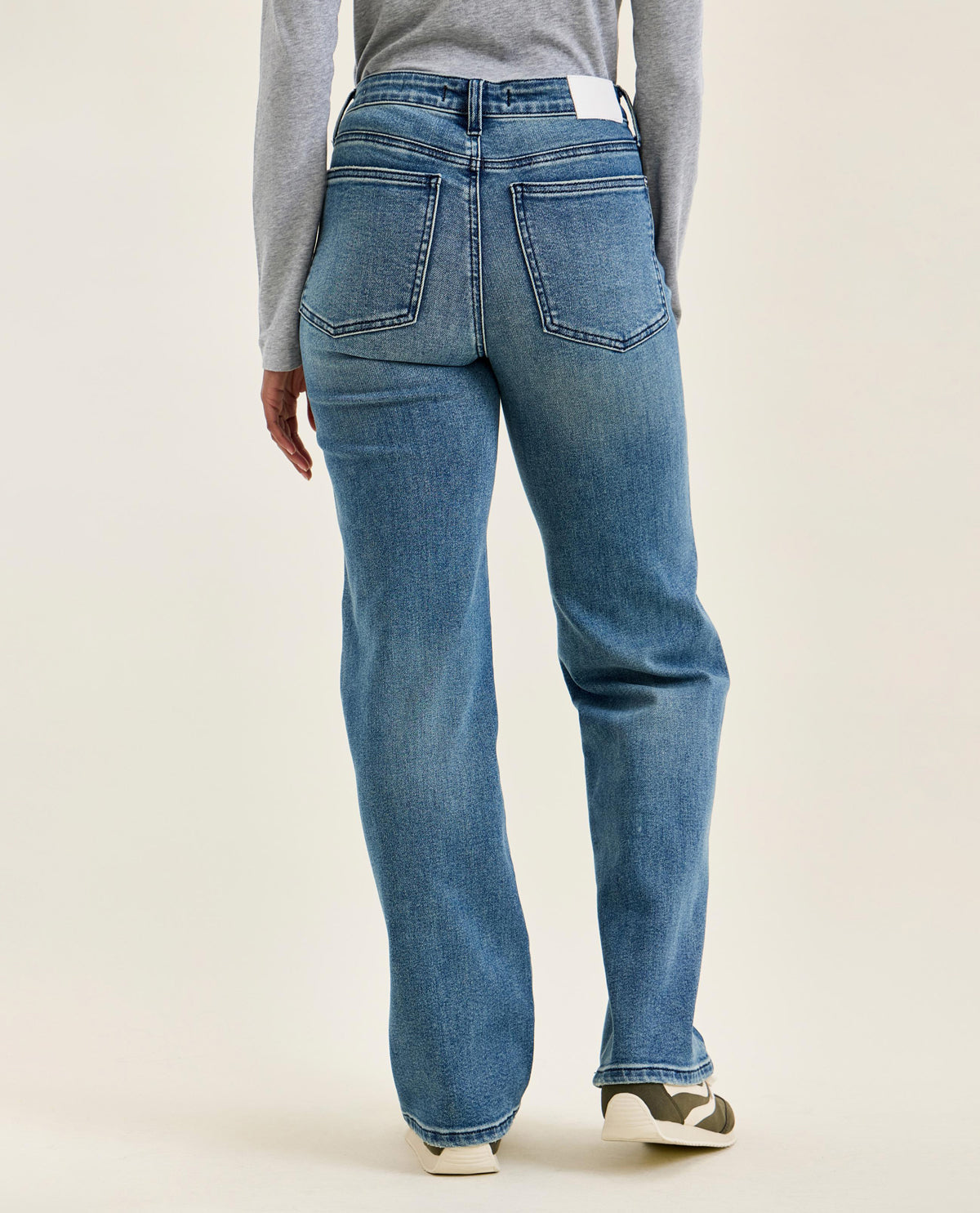 Rear view of a person wearing medium-wash blue jeans with a gray long-sleeve top and white sneakers, photographed against a plain light beige studio background.