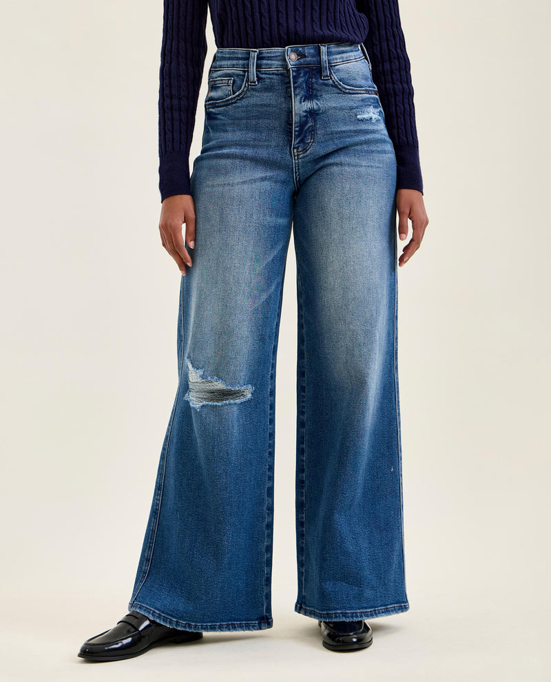 A model wearing a pair of wide-leg blue denim jeans with distressed details on the knees, paired with a dark navy ribbed sweater and black shoes, against a plain light beige studio background.