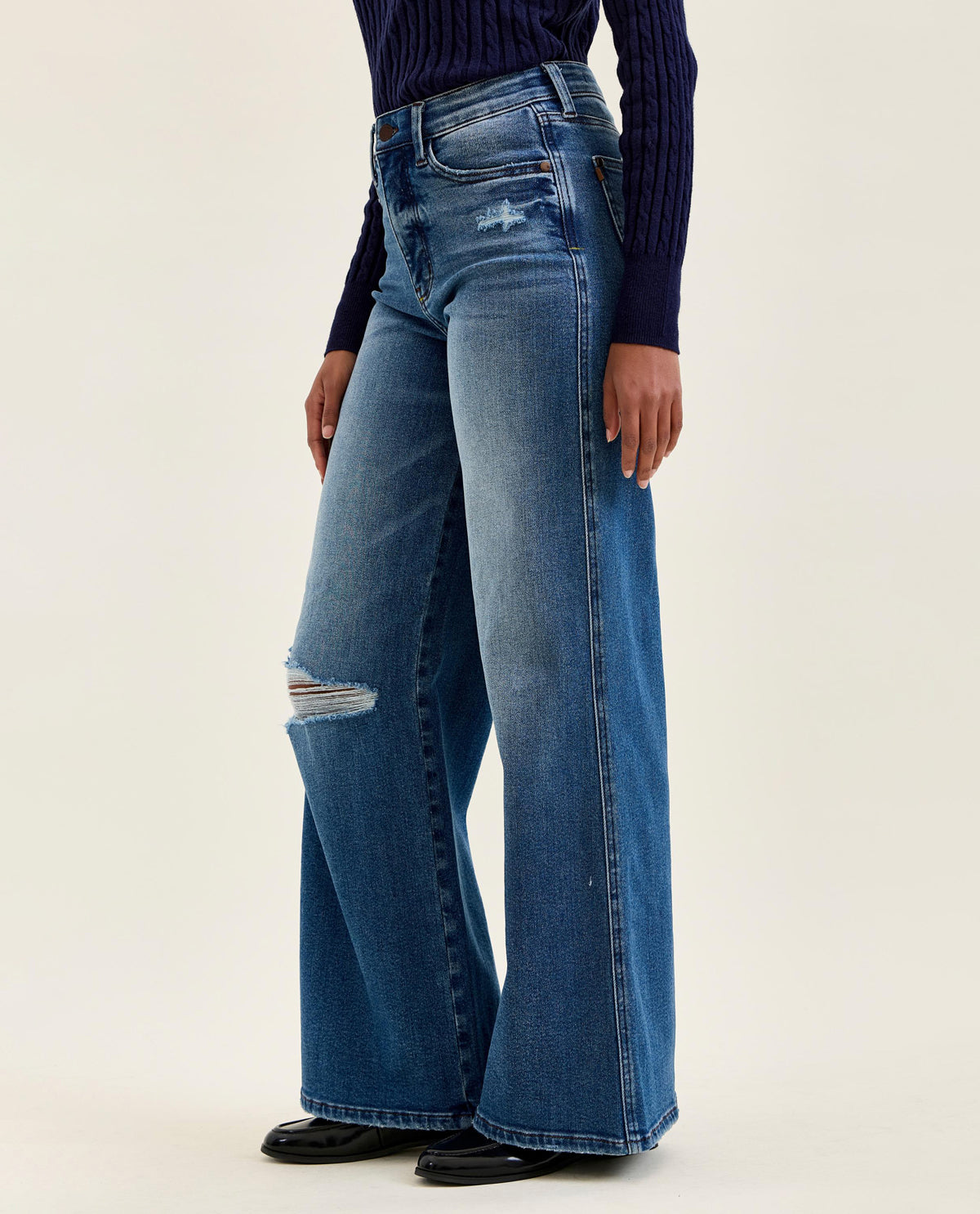 A model wearing a pair of wide-leg blue denim jeans with distressed details on the knees, paired with a dark navy ribbed sweater and black shoes, against a plain light beige studio background.