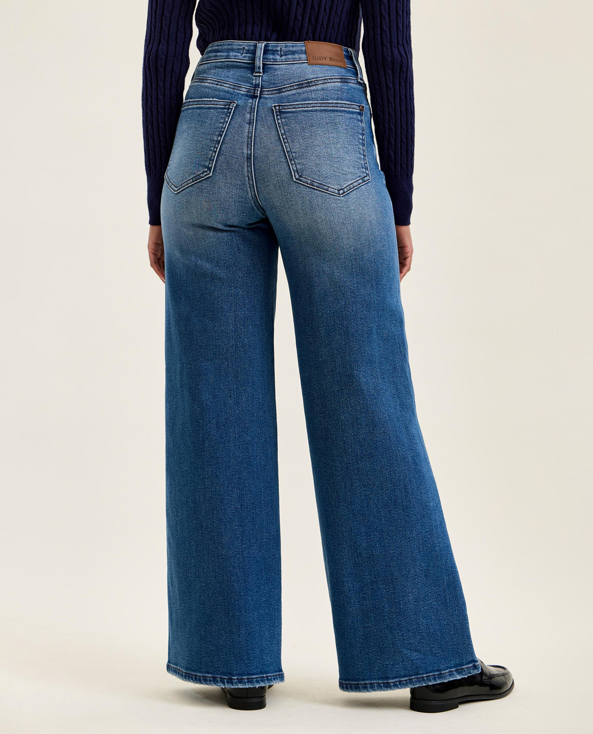 Rear view of a person wearing blue wide-leg jeans with a dark navy ribbed long-sleeve top and black shoes, photographed against a plain light beige studio background.