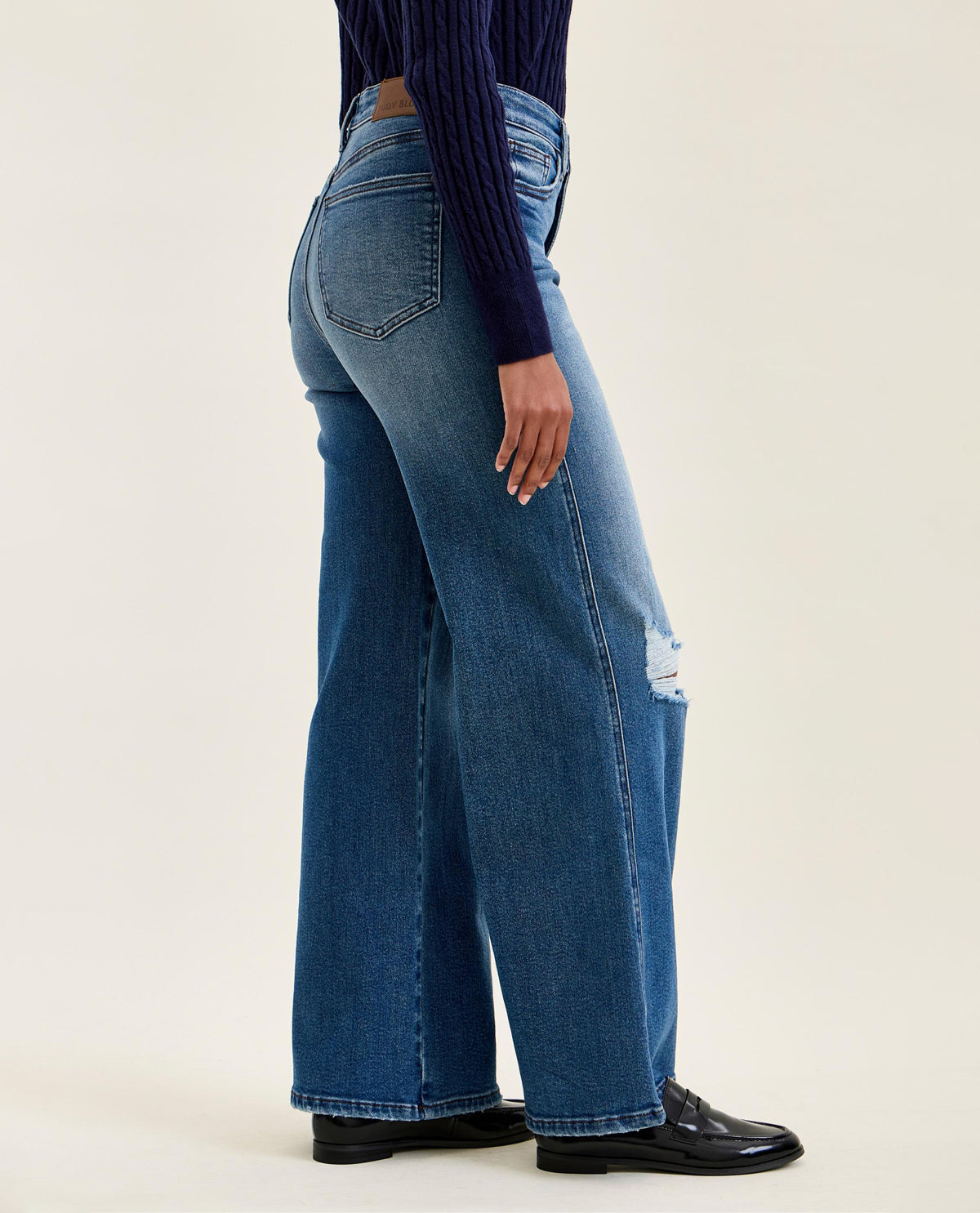A side profile view of a model wearing high-waisted, wide-leg blue jeans with a distressed rip on the upper thigh, paired with a dark blue ribbed top and black leather shoes, set against a plain light beige background.