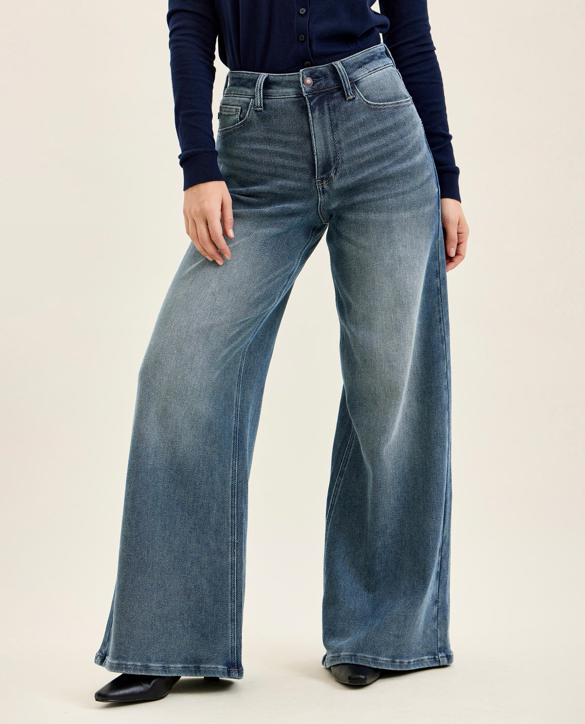 A person wearing high-waisted, wide-leg blue denim jeans with a navy long-sleeve top and black pointed shoes, photographed against a plain light beige studio background.