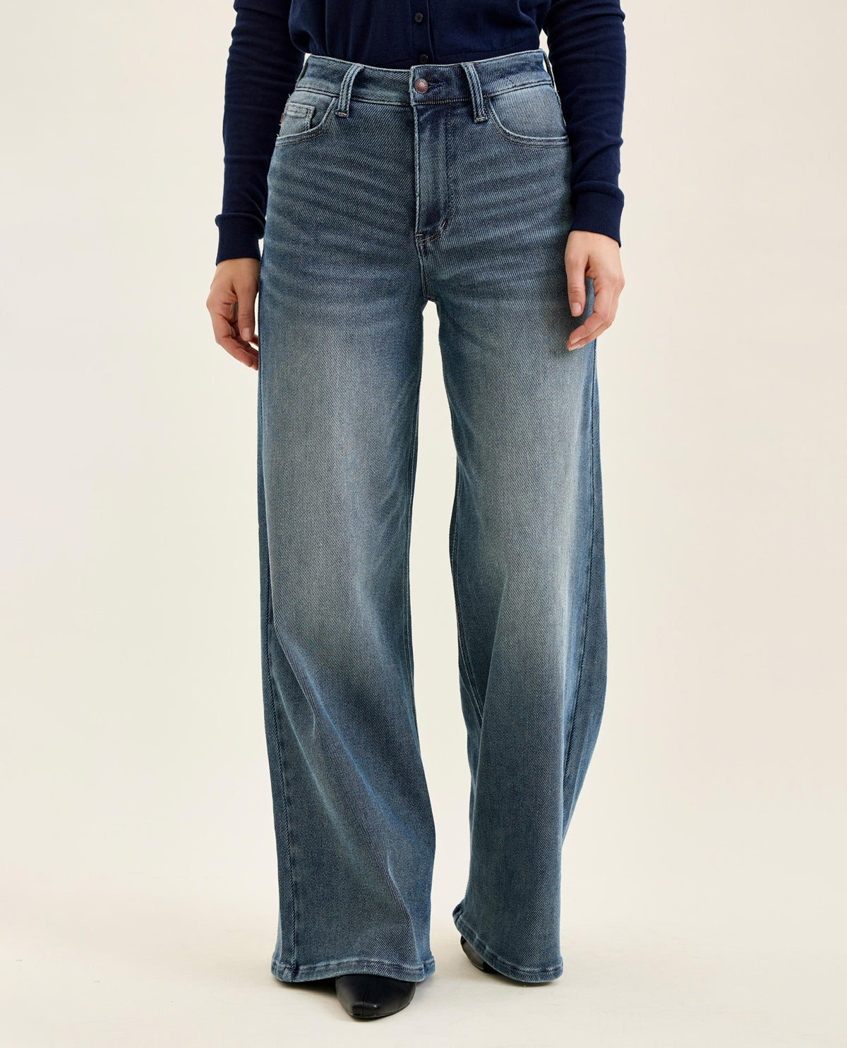 A person wearing high-waisted, wide-leg blue denim jeans with a faded wash, paired with a dark navy long-sleeve top and black shoes, against a plain light beige studio background.