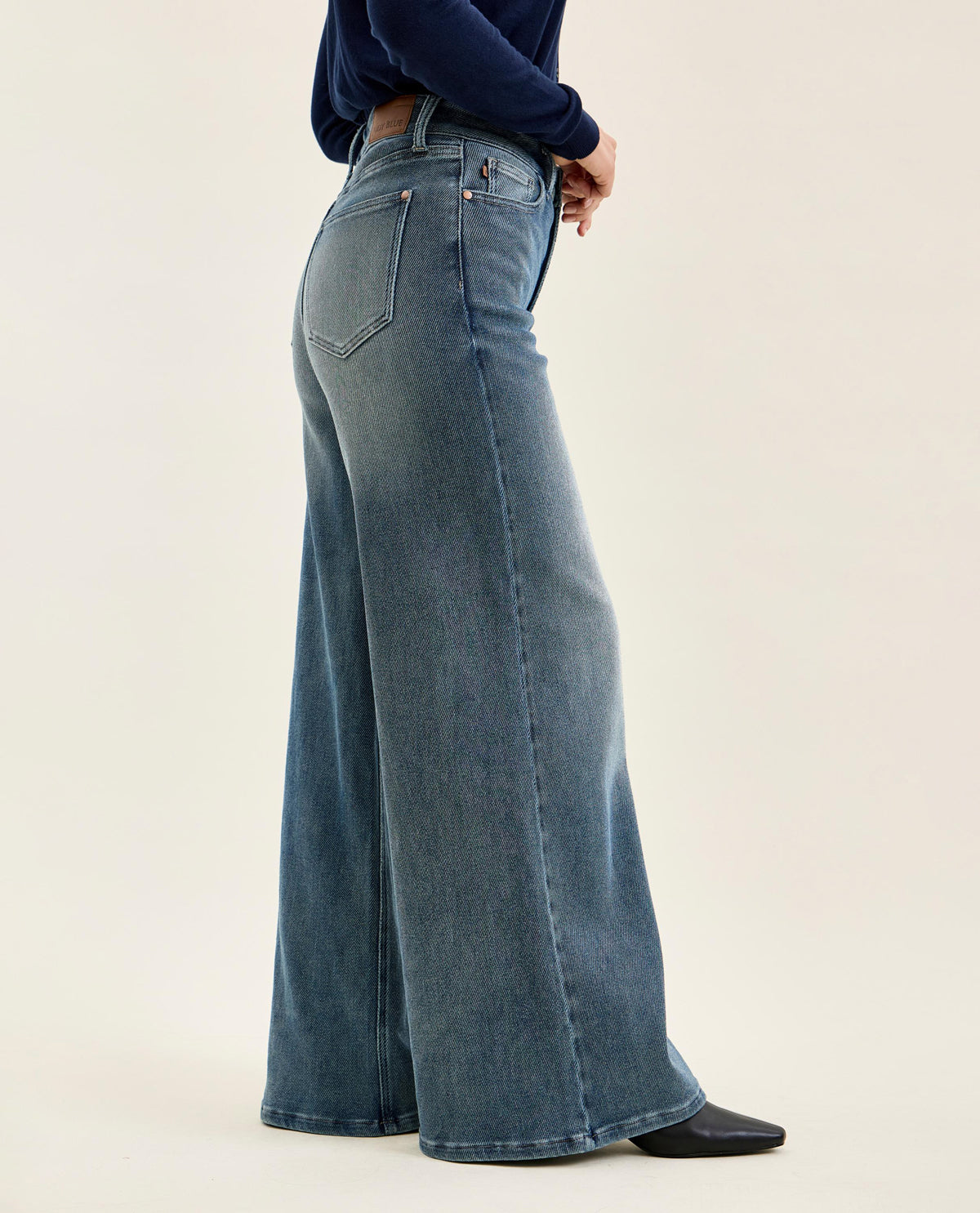 A person wearing high-waisted, wide-leg blue denim jeans with a dark navy long-sleeve top and black shoes, shown from the side against a plain light beige studio background.