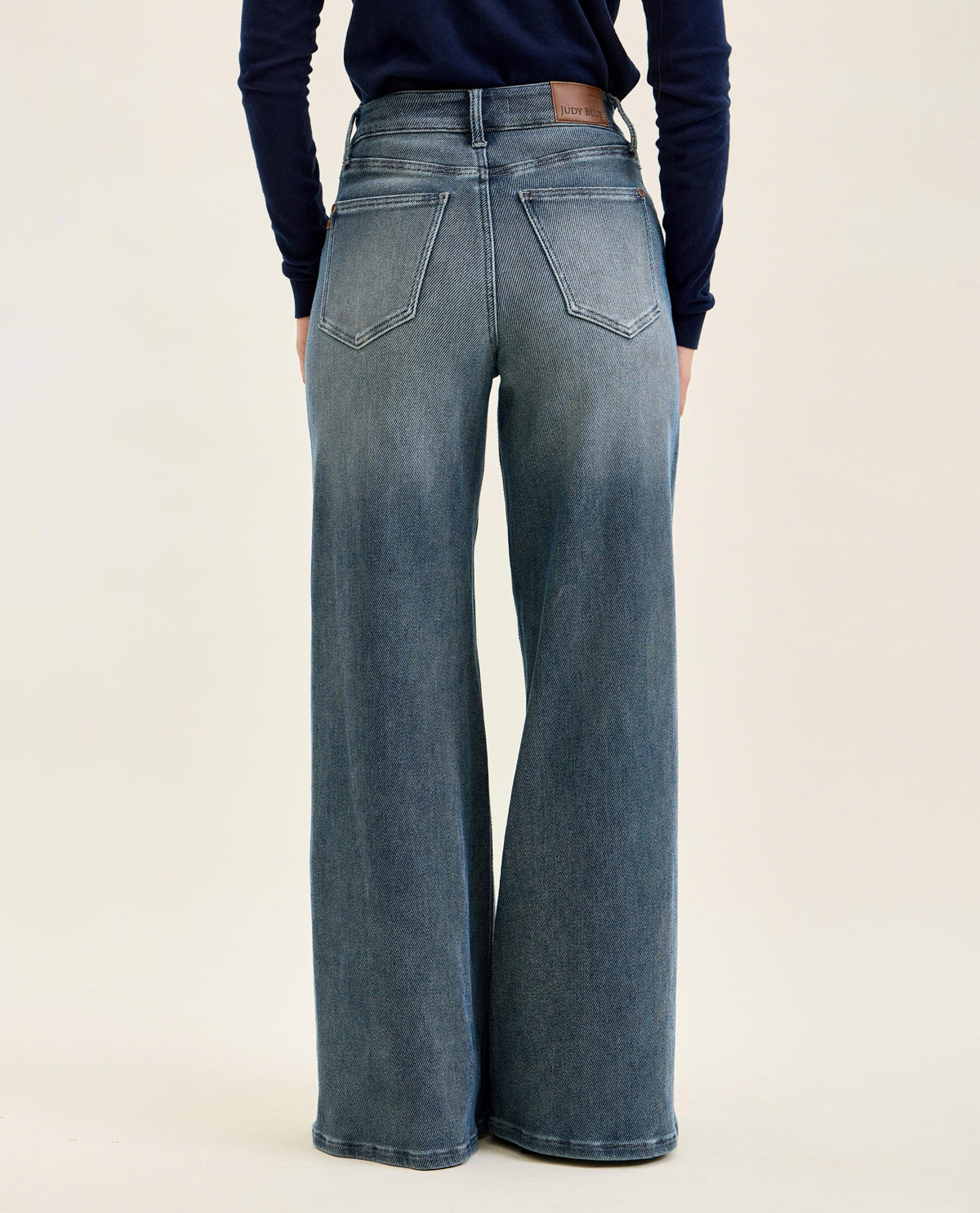 Rear view of a person wearing high-waisted, wide-leg blue denim jeans with a dark navy long-sleeve top, set against a plain light beige studio background.