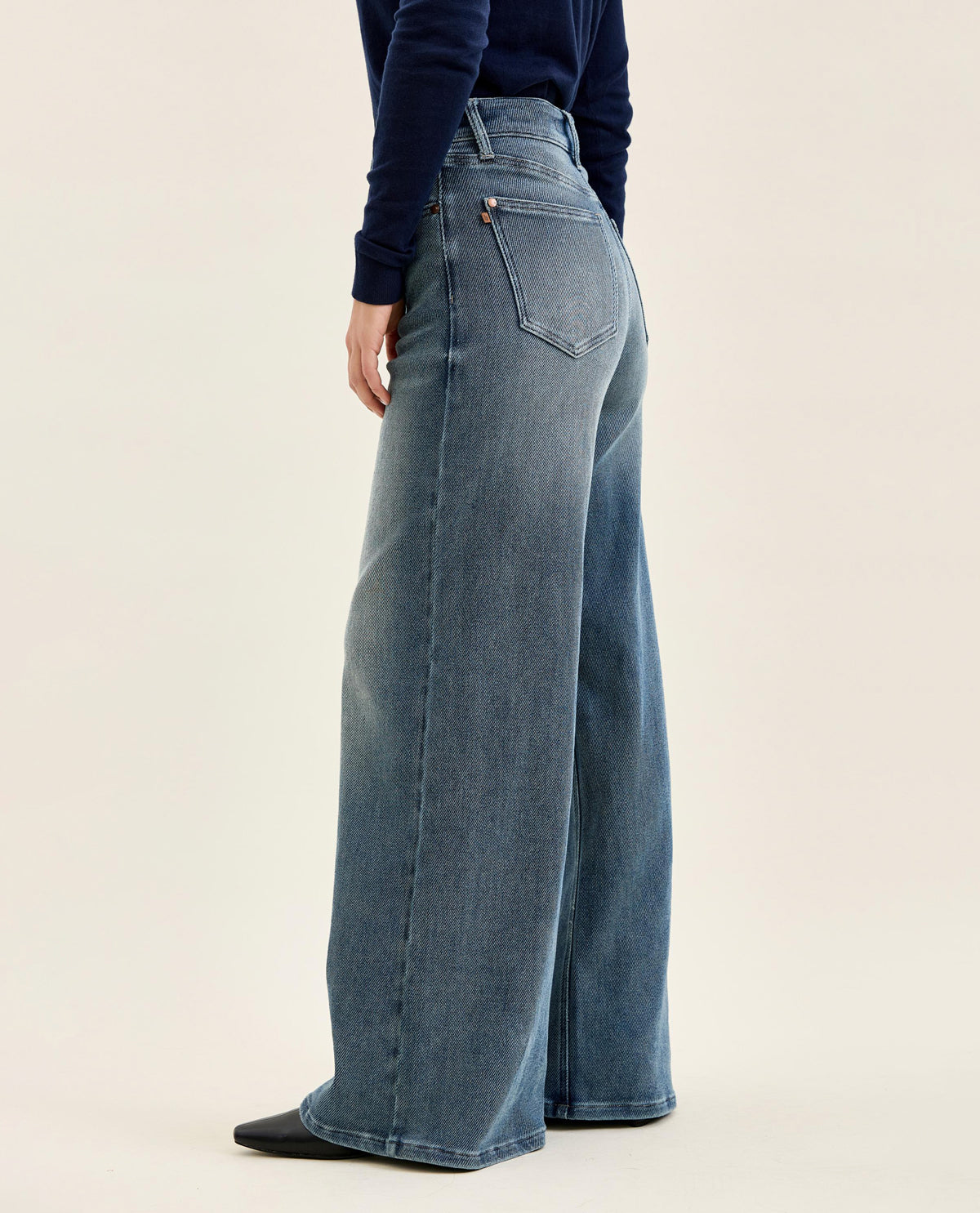 A model shown from the back wearing high-waisted, wide-leg blue denim jeans and a dark navy long-sleeve top, set against a plain light beige studio background.