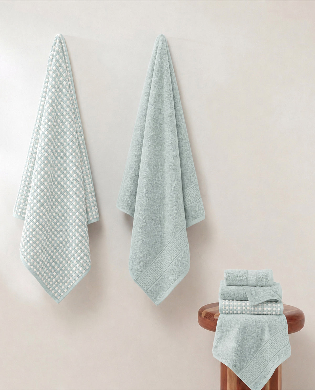 A set of pale mint green towels, including hand towels and a bath towel, displayed against a warm beige wall. One towel hangs on the left with a textured dotted pattern, while a matching folded towel set sits on a wooden stool on the right.