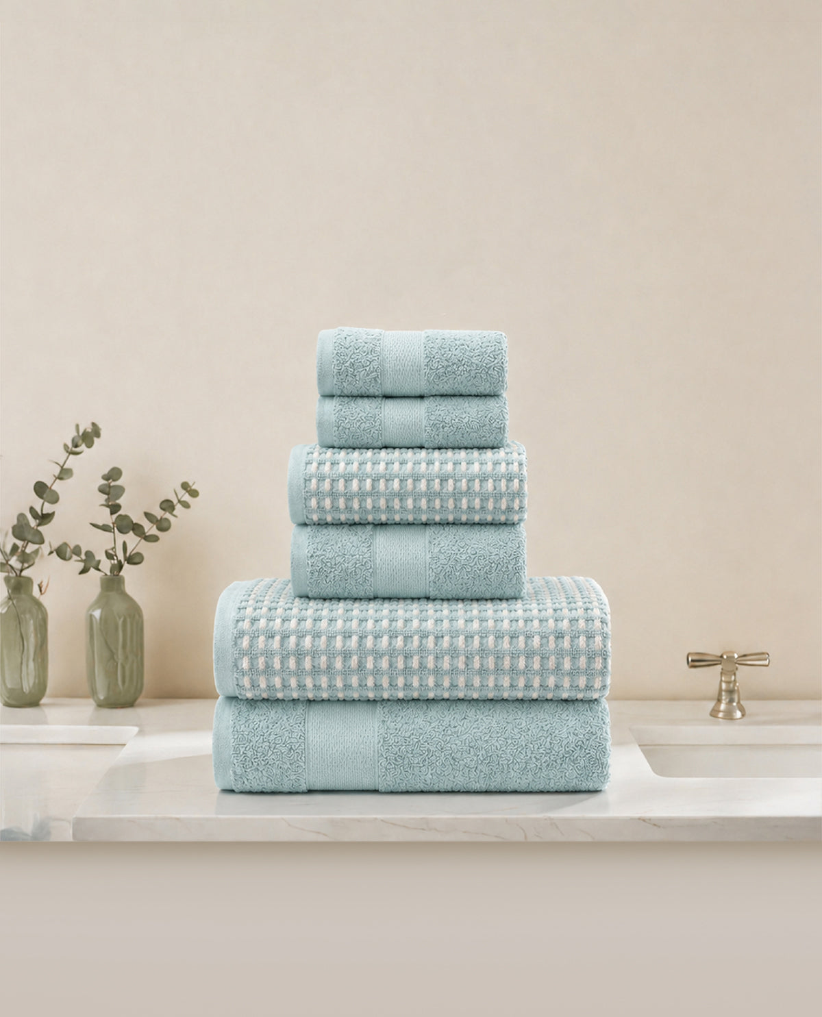 A stack of five light blue-green towels in various sizes, including two bath towels, two hand towels, and two washcloths, arranged on a white marble countertop next to a sink faucet and decorative green vases.