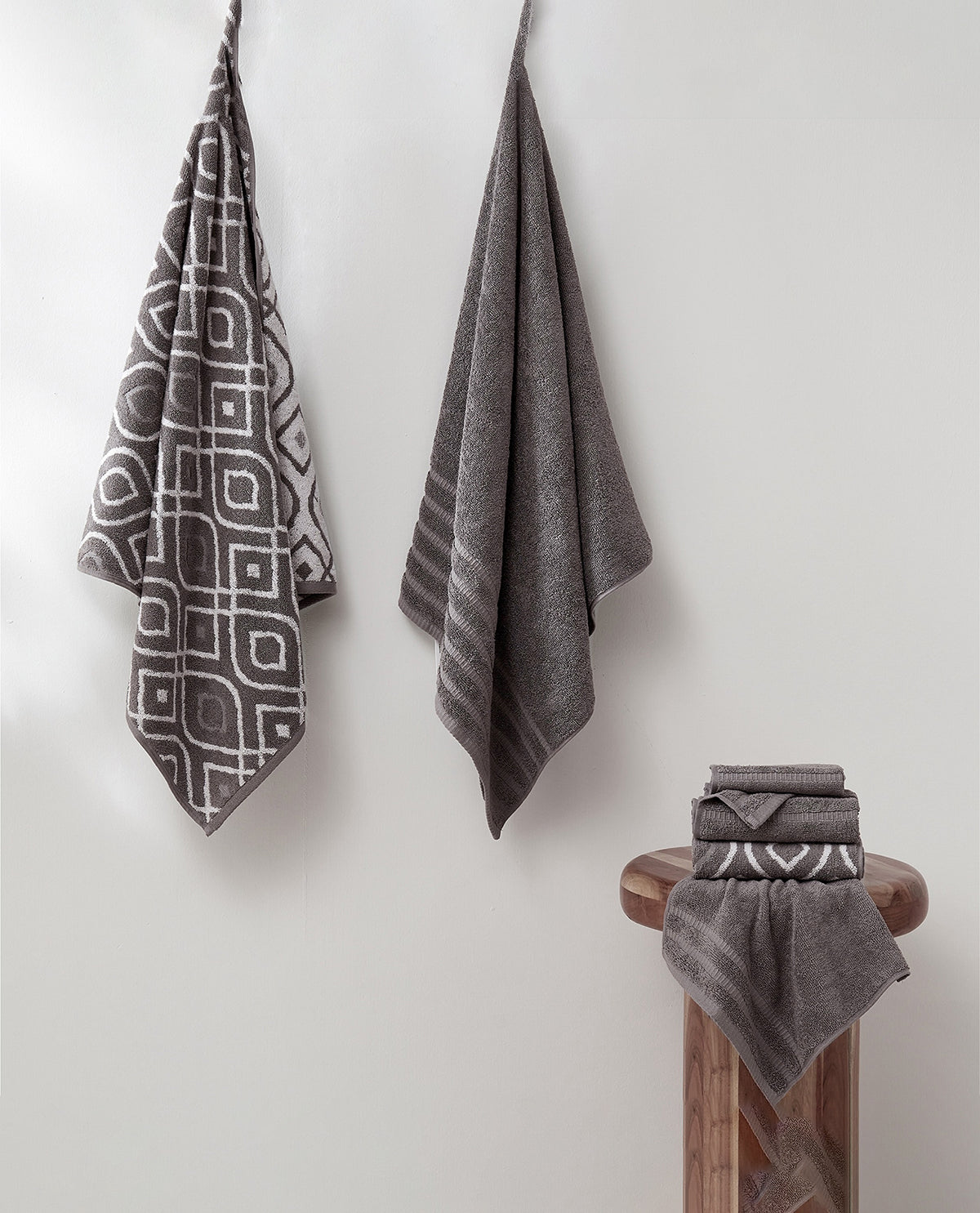 Two gray towels hanging from hooks on a light gray wall and a folded stack of two hand towels and two washcloths on a wooden pedestal.